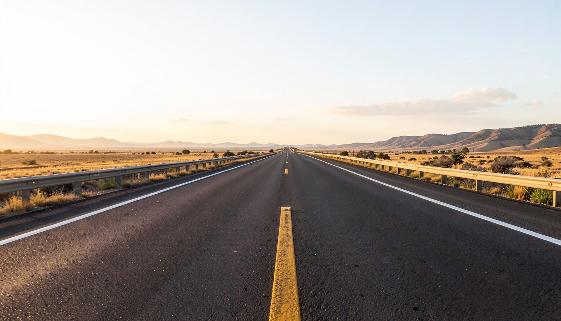 Open highway stretching toward the horizon under a bright sky, with soft golden light and distant hills